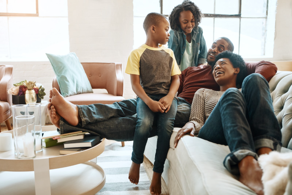 A happy family of four relaxing together on a comfortable sofa in a bright, modern living room, with two children sitting and standing near their parents. A round coffee table with books and glasses sits in the foreground.